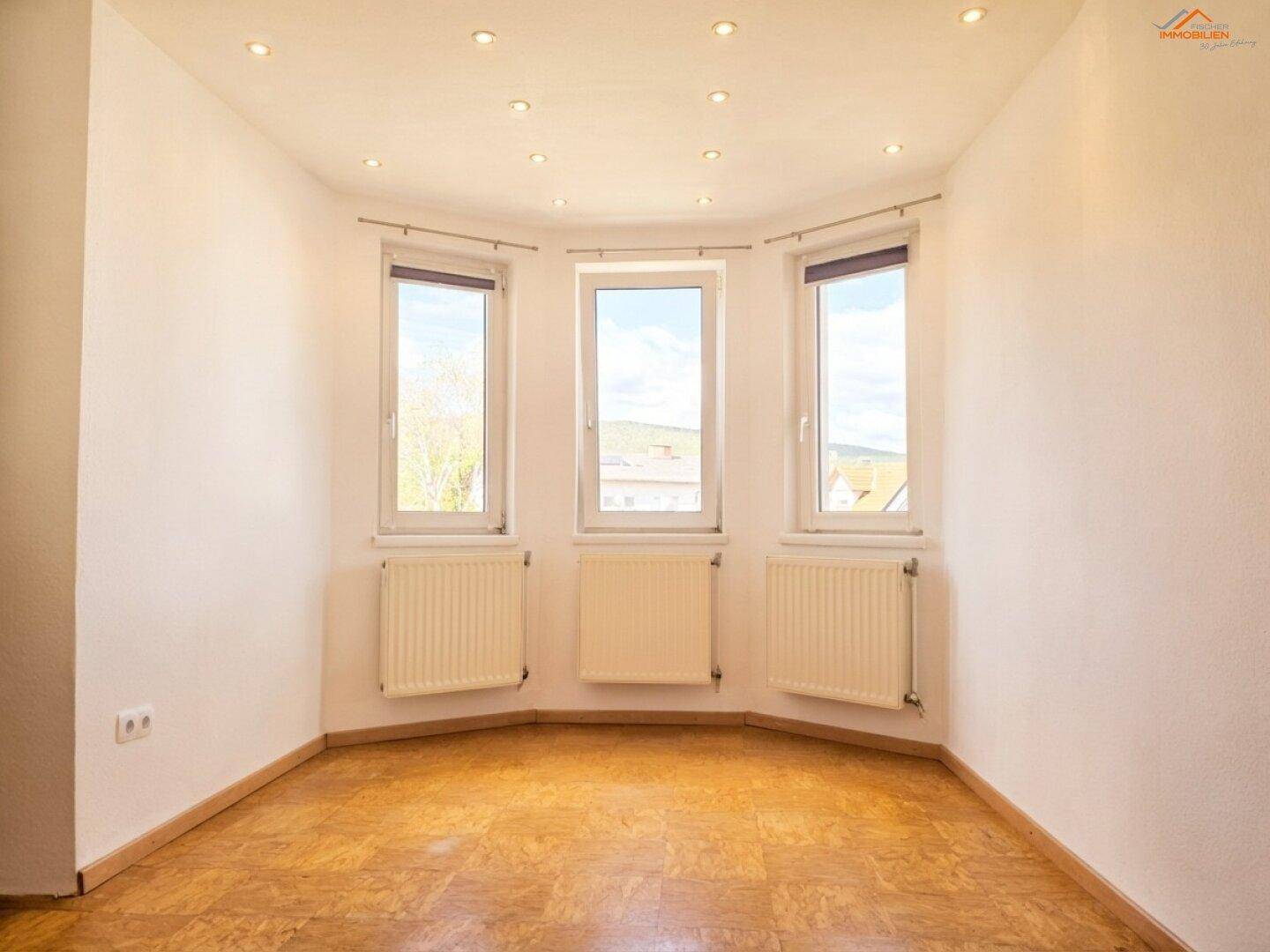 Helles Zimmer mit Erkerfenstern, Parkettboden und Einbaustrahlern an der Decke.