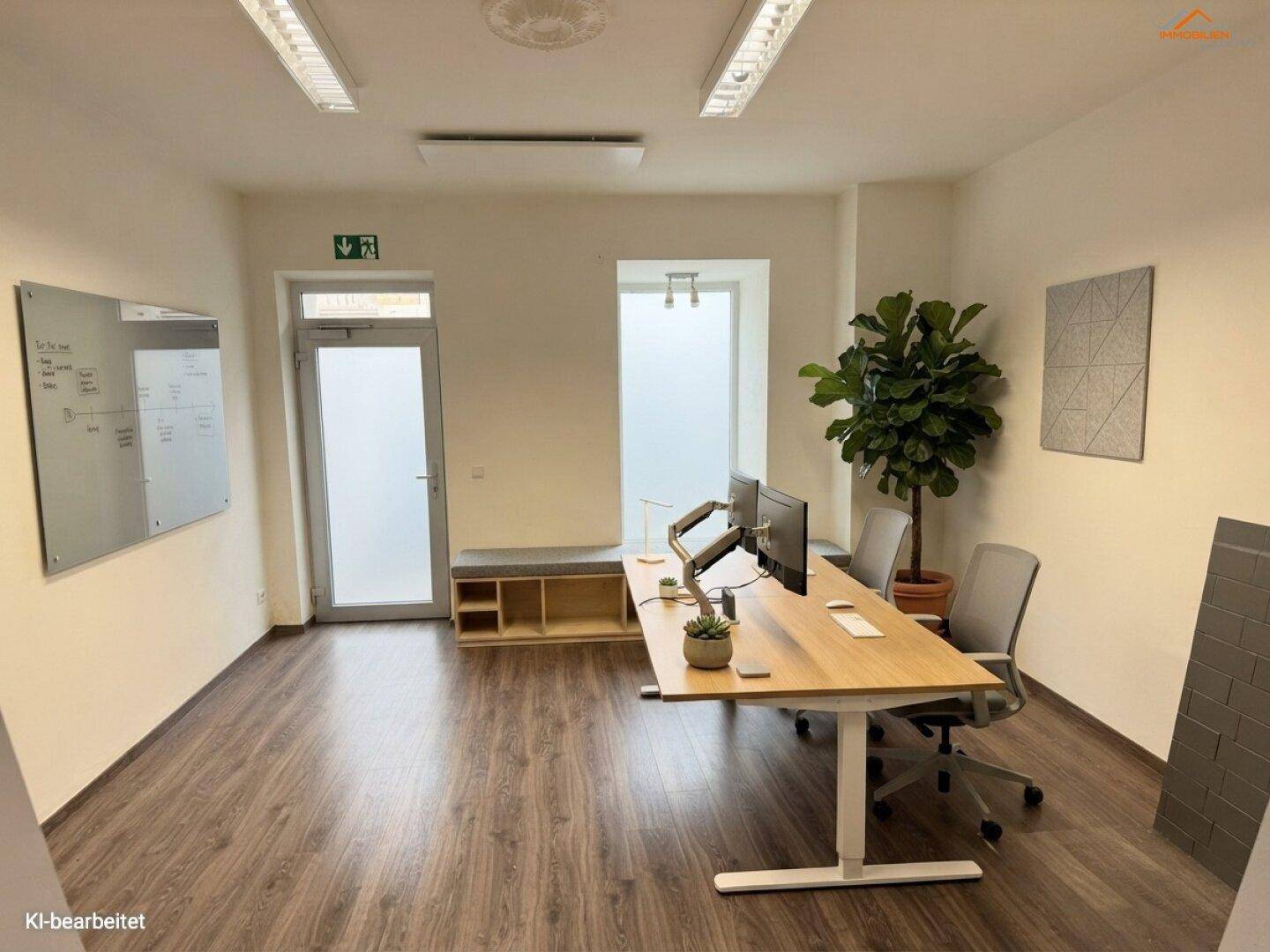 Heller Büroraum mit zwei Arbeitsplätzen, großen Fenstern und einer Sitzbank am Eingangsbereich.