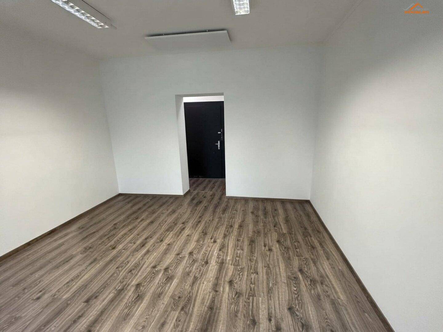 Leerer Büroraum mit Laminatboden, weißen Wänden und einer dunklen Tür im Hintergrund.