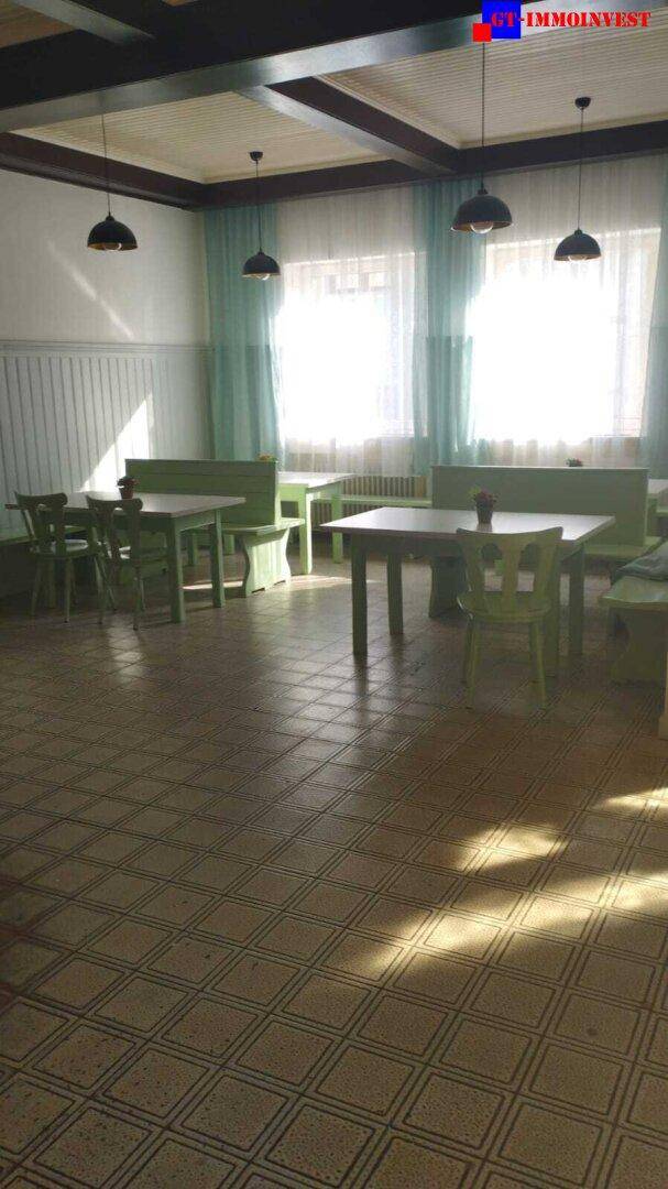 Essbereich mit grünen Sitzbänken und Tischen, großen Fenstern mit Vorhängen und gefliestem Boden.