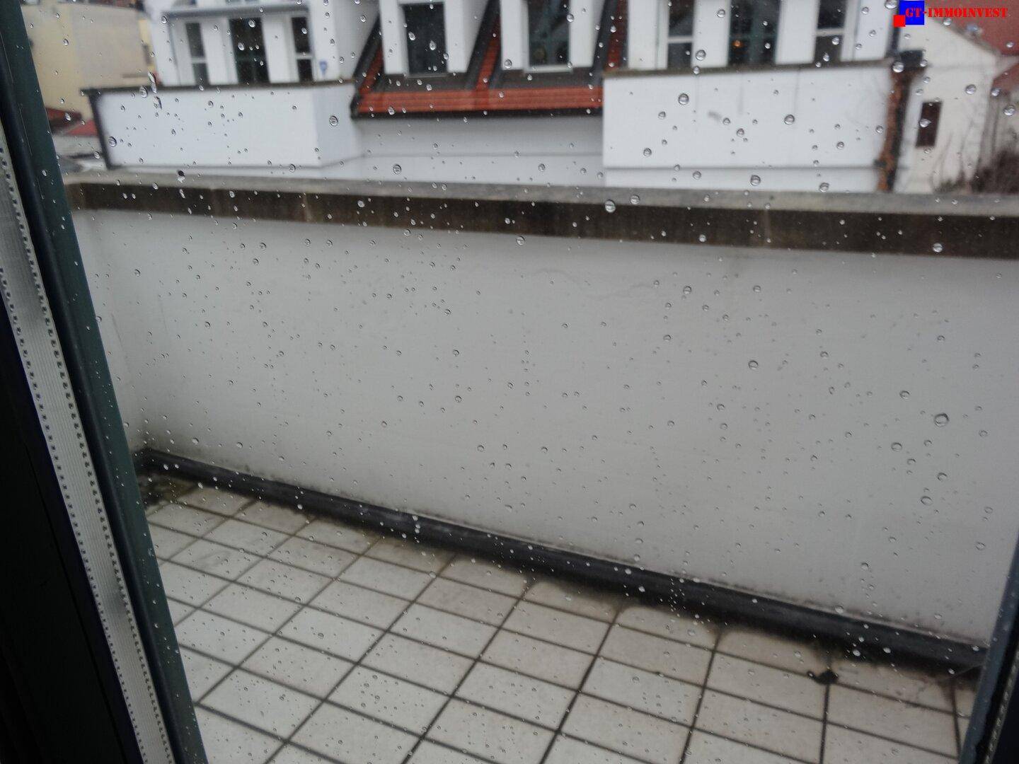 Balkon mit Fliesenboden und Blick auf umliegende Gebäude, sichtbar durch ein regennasses Fenster.