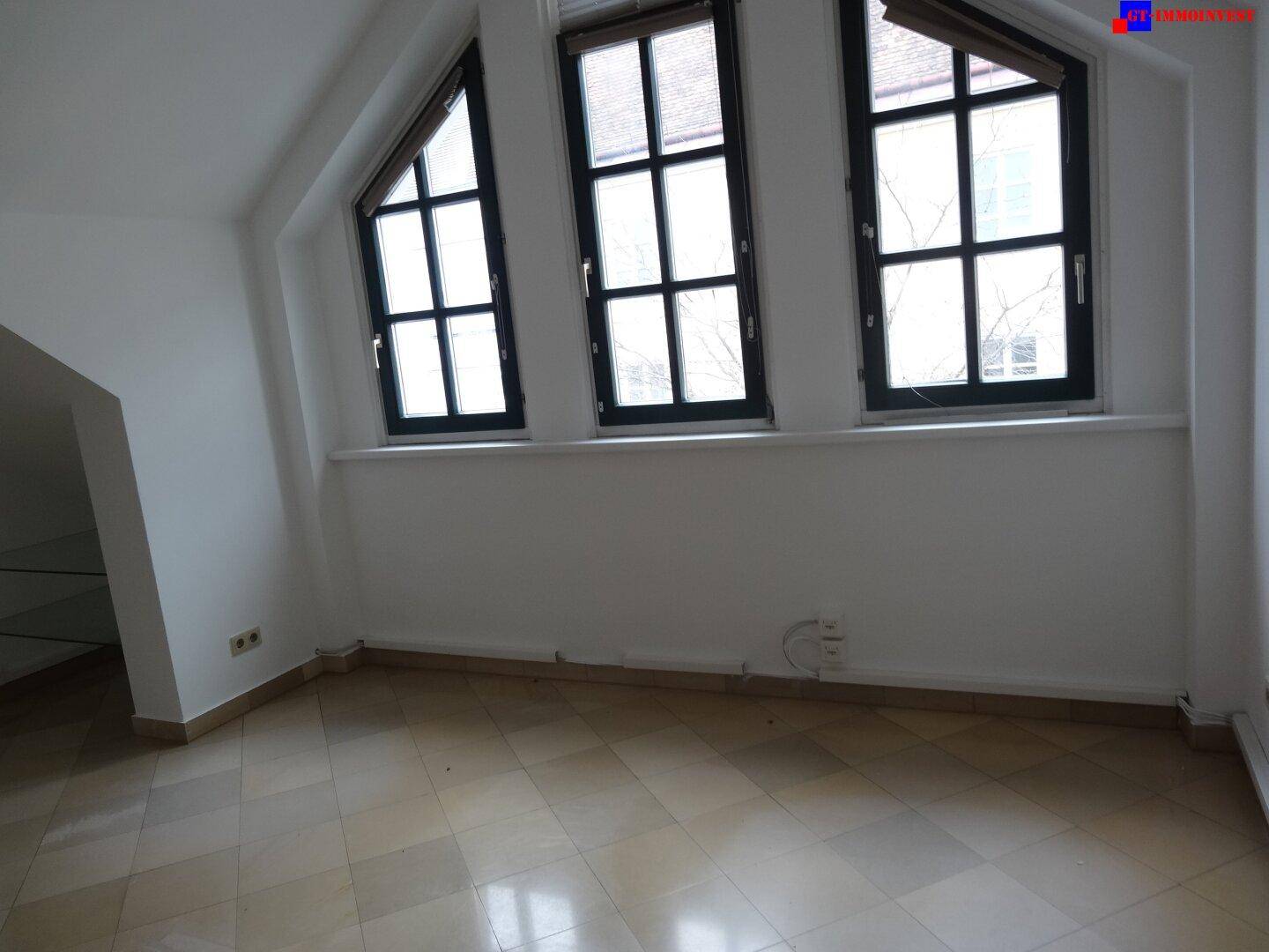 Wohnbereich mit drei hohen Fenstern, die viel Tageslicht hereinlassen und den Raum hell erscheinen lassen.