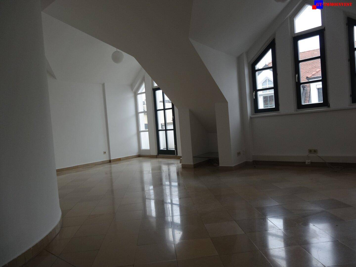 Heller Wohnbereich mit schrägen Wänden, großen Fenstern und glänzenden Fliesenböden, die den Raum offen wirken lassen.