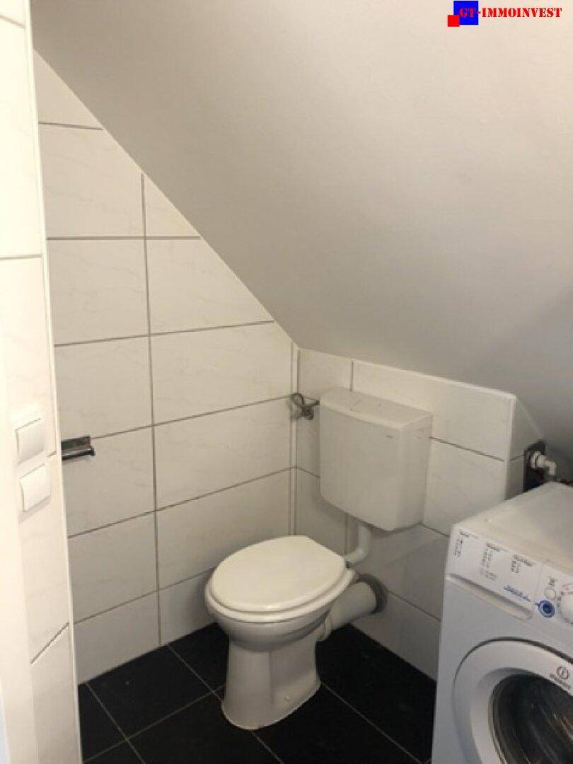 WC im Dachgeschoss mit weißen Fliesen, Toilette und Platz für eine Waschmaschine.