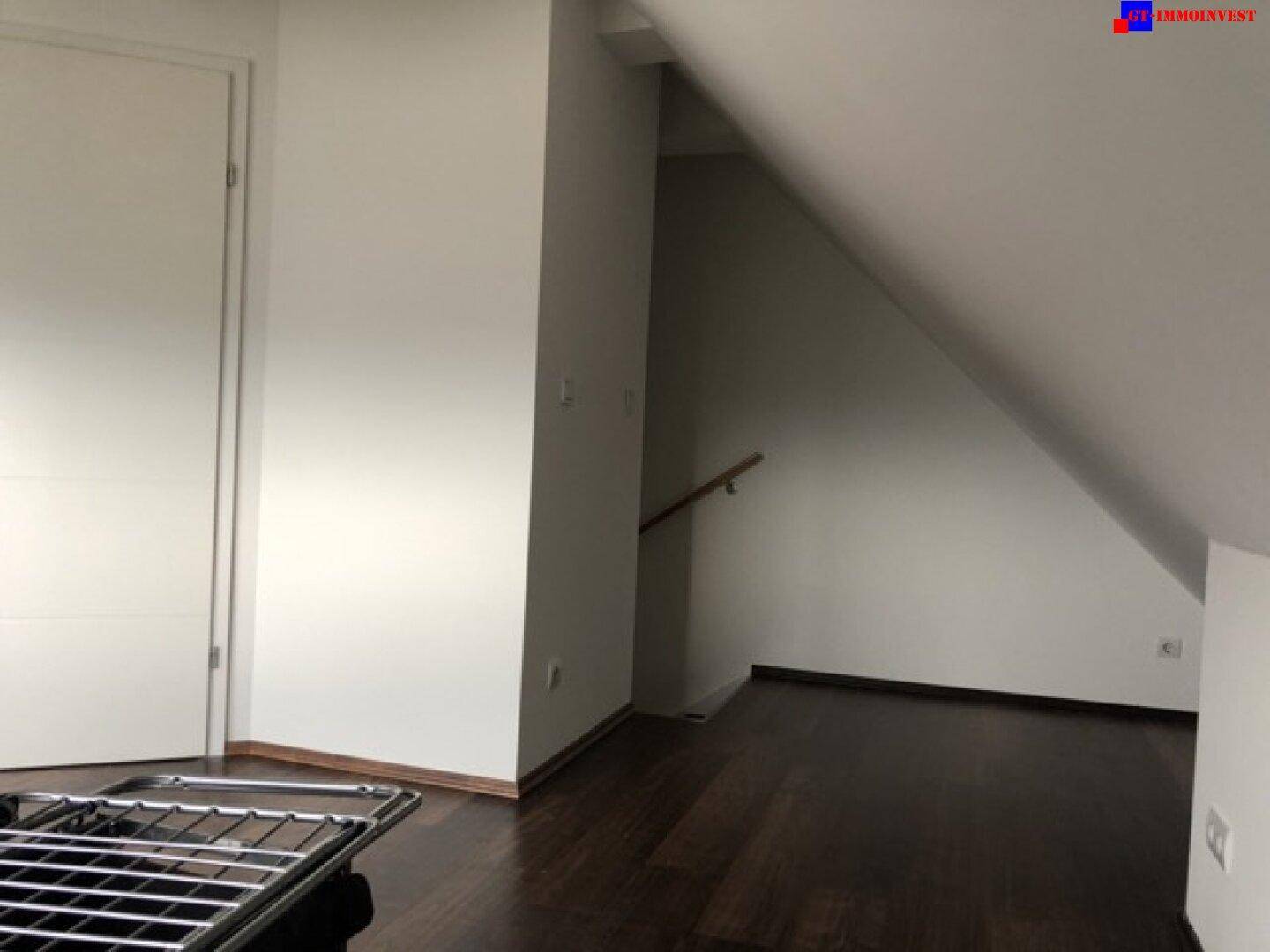 Dachgeschossraum mit dunklem Holzboden, weiß gestrichenen Wänden und Zugang zu einem weiteren Bereich.