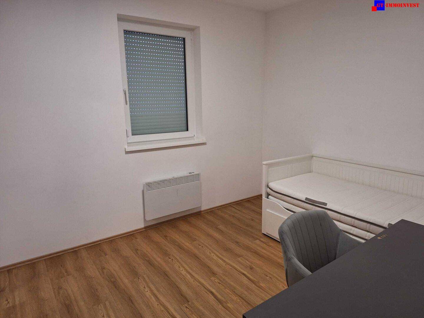 Helles Schlafzimmer mit Einzelbett, Laminatboden und Fenster mit Rollladen.