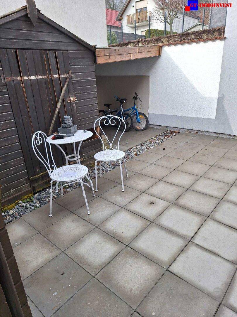 Gepflasterter Terrassenbereich mit Gartenmöbeln und einem Holzschuppen im Hintergrund.