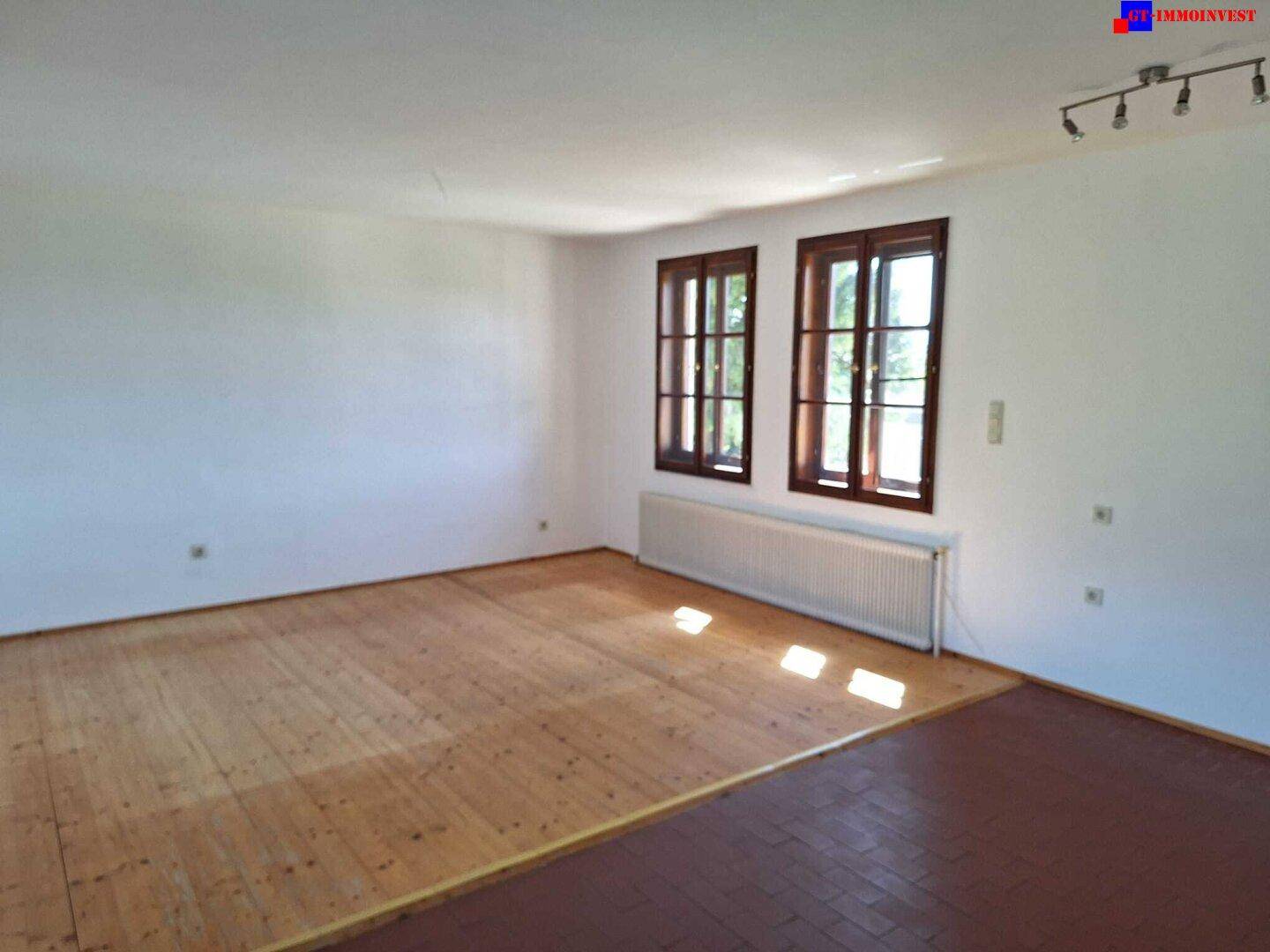 Heller Wohnraum mit Holzboden, zwei Fenstern und einem Heizkörper unter blauem Himmel.