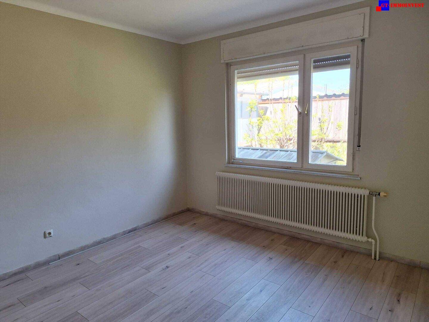 Schlafzimmer mit hellem Laminatboden, einem Fenster mit Rollladen und einem Heizkörper.