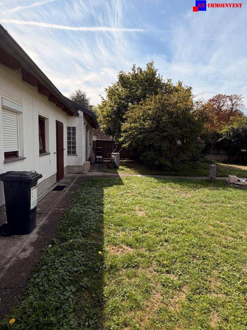Seitenansicht des Hauses mit einem gepflasterten Weg, Rasenfläche und üppigem Grün im Hintergrund.