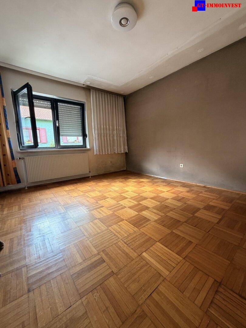Schlafzimmer mit Parkettboden, schlichten Wänden und einem Fenster mit Heizkörper darunter.