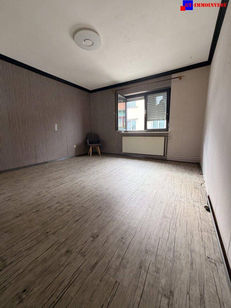 Zimmer mit Laminatboden, gestreifter Tapete und einem Fenster mit Heizkörper.