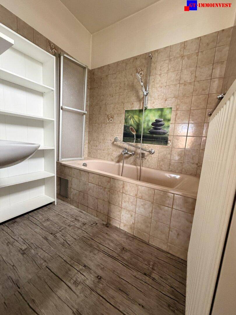 Badezimmer mit beigefarbenen Fliesen, einer Badewanne mit Duschfunktion und einem Waschbecken mit Regal.