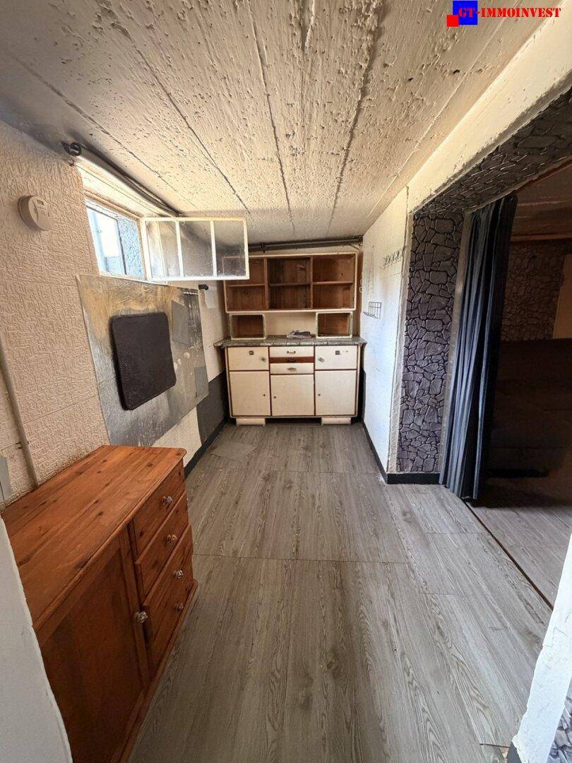 Kellerraum mit alter Spüle und Holzkommode, die Wände sind mit strukturierter Tapete versehen.