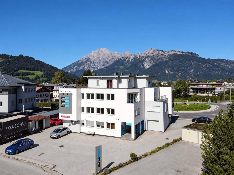 Ansicht des modernen Bürogebäudes mit Parkplätzen und majestätischen Bergen im Hintergrund an einem sonnigen Tag.