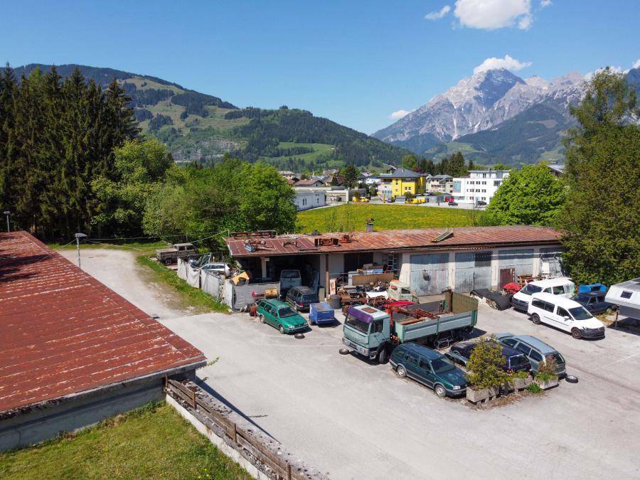 Ansicht der Gebäude und des Parkplatzes mit Fahrzeugen, umgeben von Bäumen und Bergen im Hintergrund.