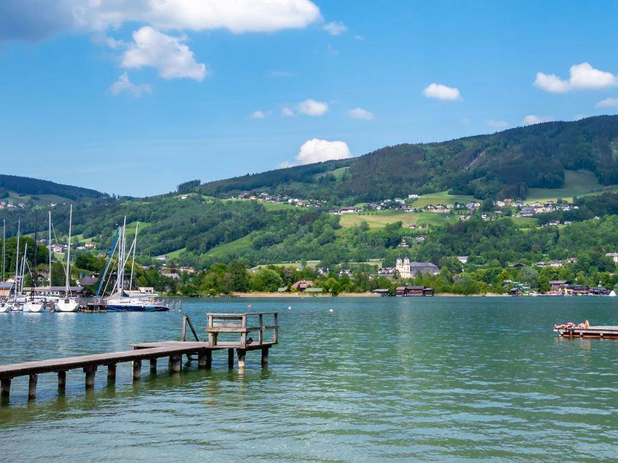 Malerischer Blick auf den Mondsee mit Segelbooten, einem Holzsteg und bewaldeten Hügeln.