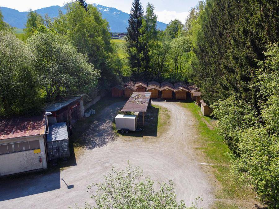 Luftaufnahme eines unbefestigten Bereichs mit kleinen Lagerhallen oder Schuppen, umgeben von Bäumen.