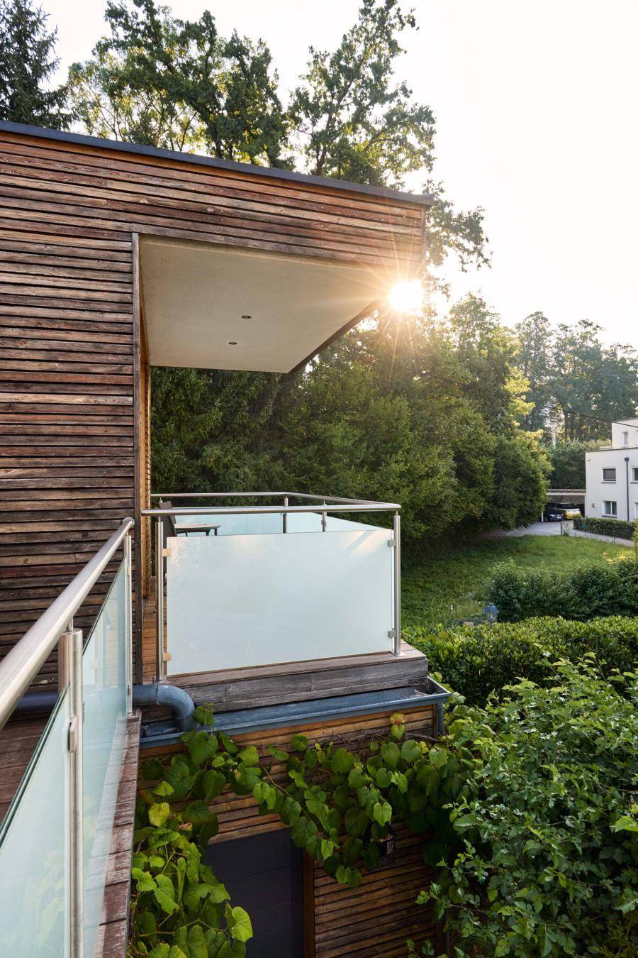 Moderner Balkon mit Glasgeländer und Holzfassade, umgeben von üppigem Grün und Sonnenschein.