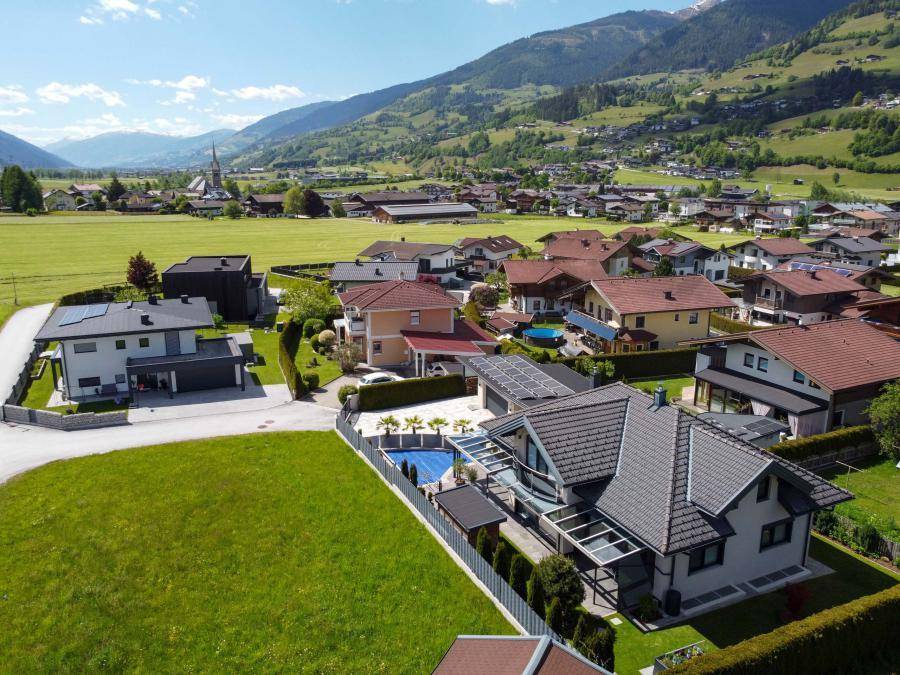 Luftaufnahme einer Wohnsiedlung mit Einfamilienhäusern und grünen Wiesen vor Bergkulisse.