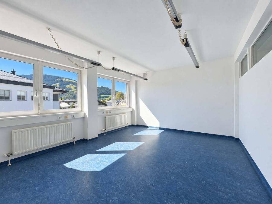Geräumiges Büro mit blauem Linoleumboden, großen Fenstern und Heizkörpern, bietet viel Tageslicht.