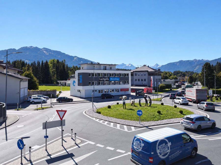 Bürogebäude an einem belebten Kreisverkehr mit Bergkulisse, umgeben von grüner Landschaft und klarem Himmel.