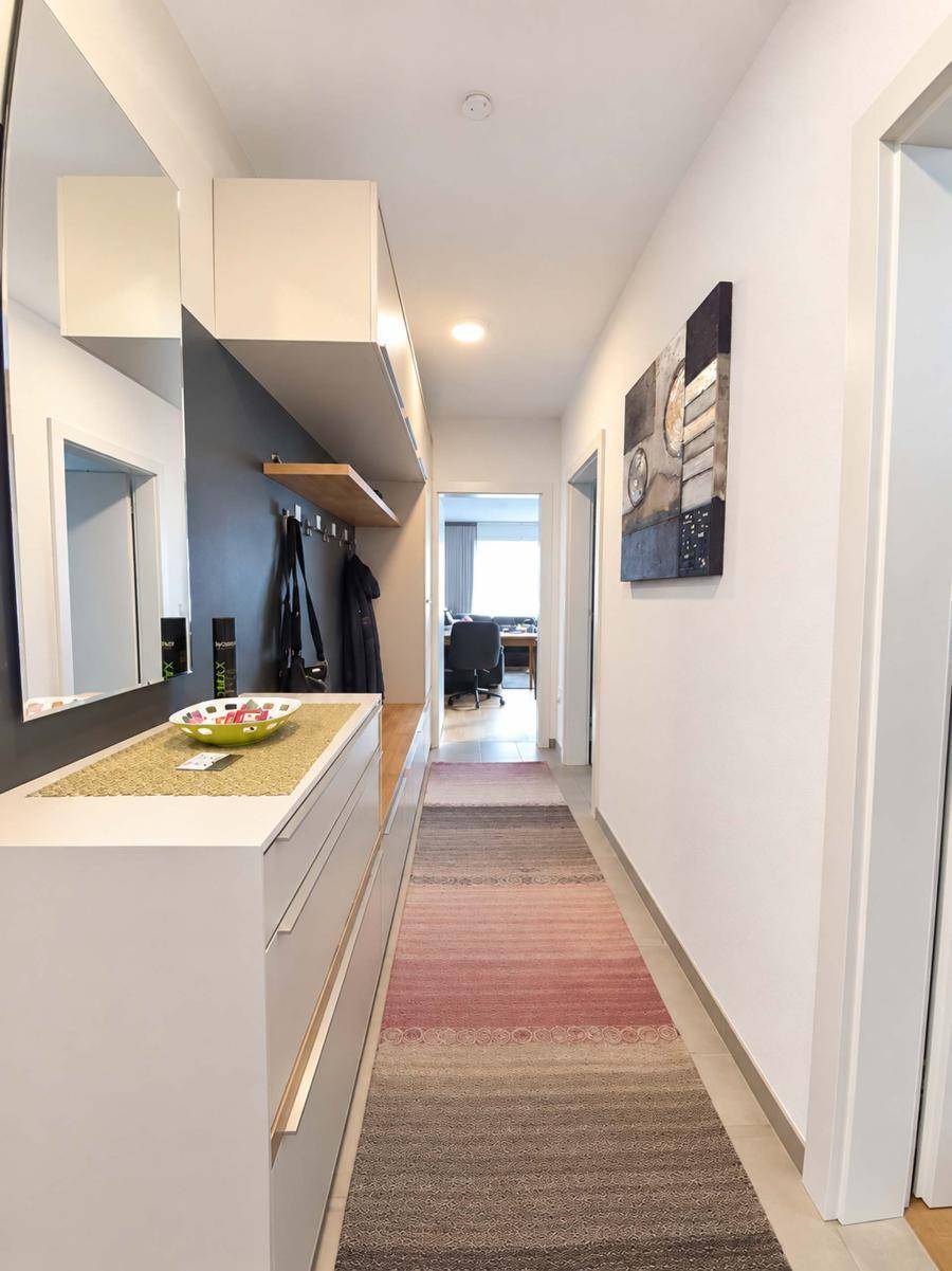 Langer, heller Flur mit modernem Teppichläufer, Garderobe und Spiegel, der zu weiteren Räumen führt.