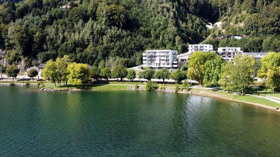Panoramablick auf den See mit grünen Ufern und modernen Gebäuden, eingebettet in die bewaldete Hügellandschaft.