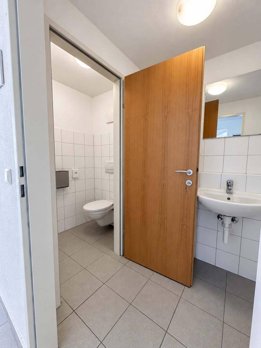 Helles Badezimmer mit weißen Fliesen, Waschbecken und Toilette, ausgestattet mit einer Holztür.