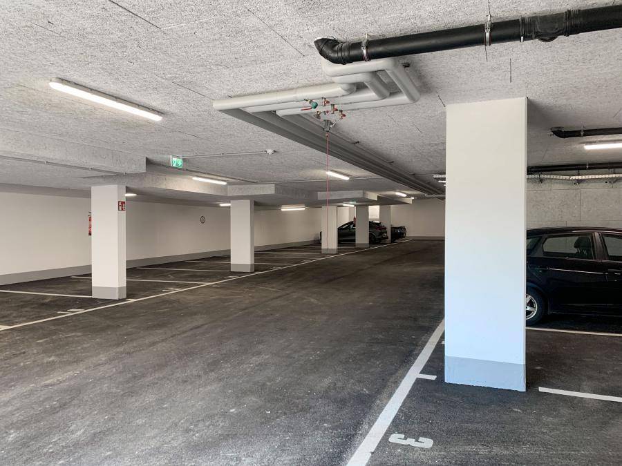 Moderne Tiefgarage mit sichtbaren Deckeninstallationen, vielen Stellplätzen und Säulenstruktur.