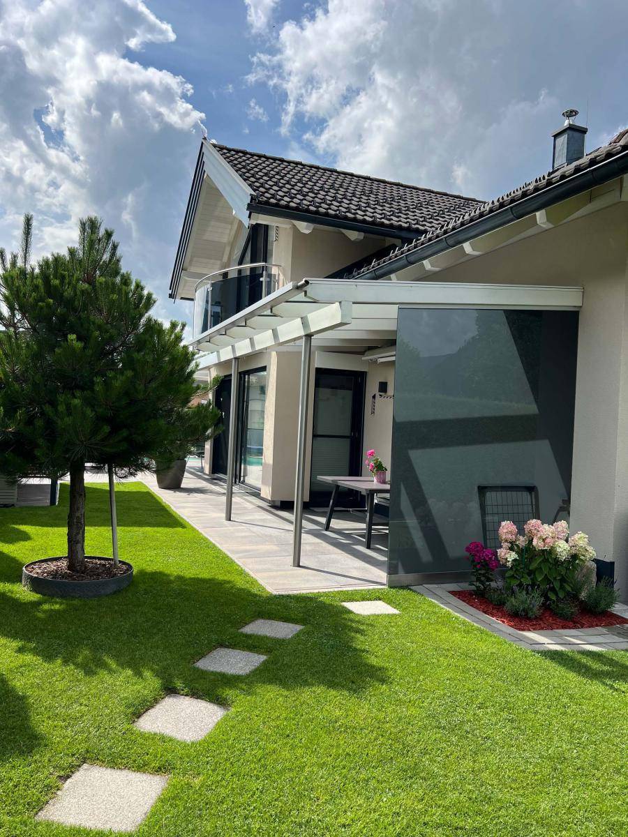 Seitlicher Blick auf das Haus mit überdachter Terrasse, kleinem Garten und Balkon im Obergeschoss.