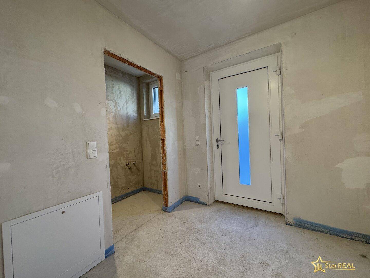 Eingangsbereich im Rohbau mit neuer Tür und Zugang zu einem kleinen Raum.