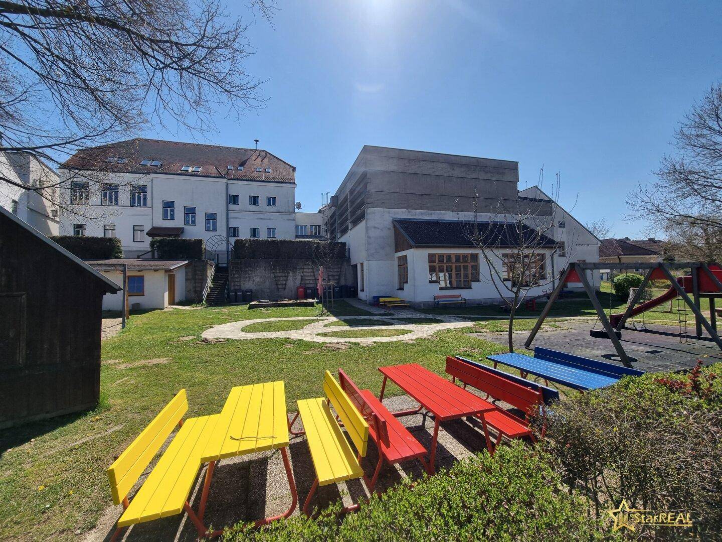 Bunter Spielplatz mit Tischen, Bänken, Schaukeln und Rutschen vor Wohngebäuden.