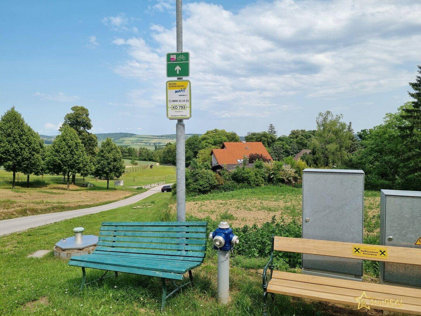 Ländliche Szene mit Sitzbänken, Hydrant und Wegweiser, umgeben von grüner Landschaft.