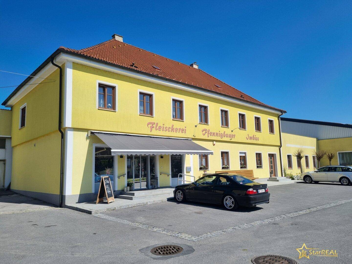 Gelbes Gebäude mit rotem Ziegeldach und Geschäftsfassade, davor ein Parkplatz.