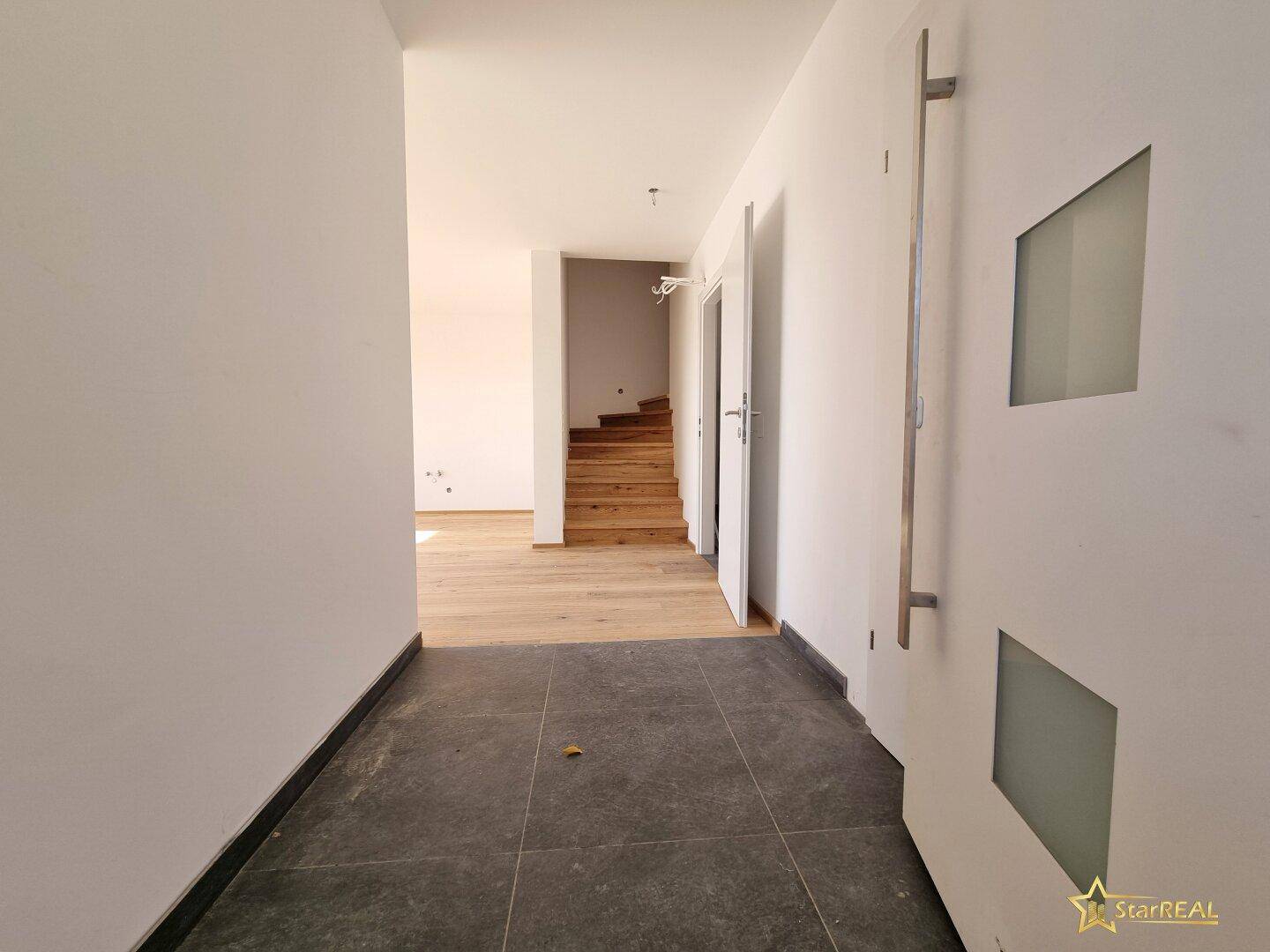 Heller Eingangsbereich mit dunklen Fliesen und einer Holztreppe, die in die oberen Stockwerke führt.
