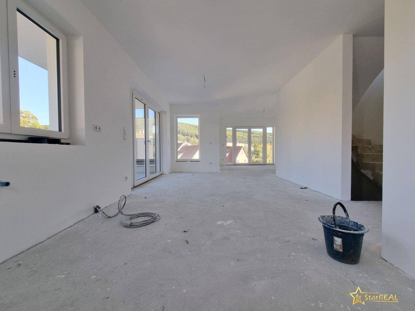Geräumiger Wohnbereich im Rohbau mit Treppenaufgang und viel Tageslicht durch große Fenster.