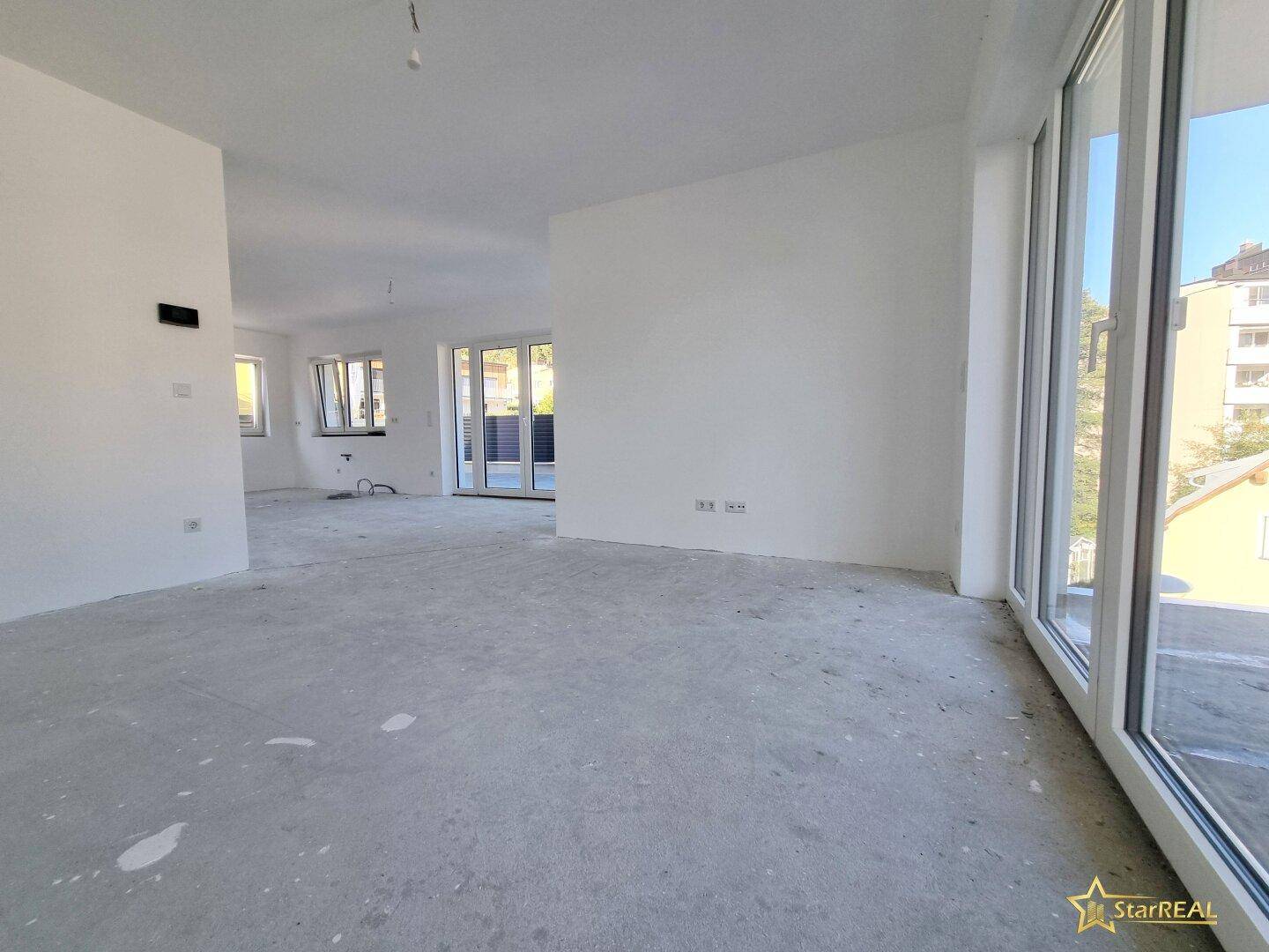 Heller Raum im Rohbau mit großen Fenstern, die viel Tageslicht hereinlassen.