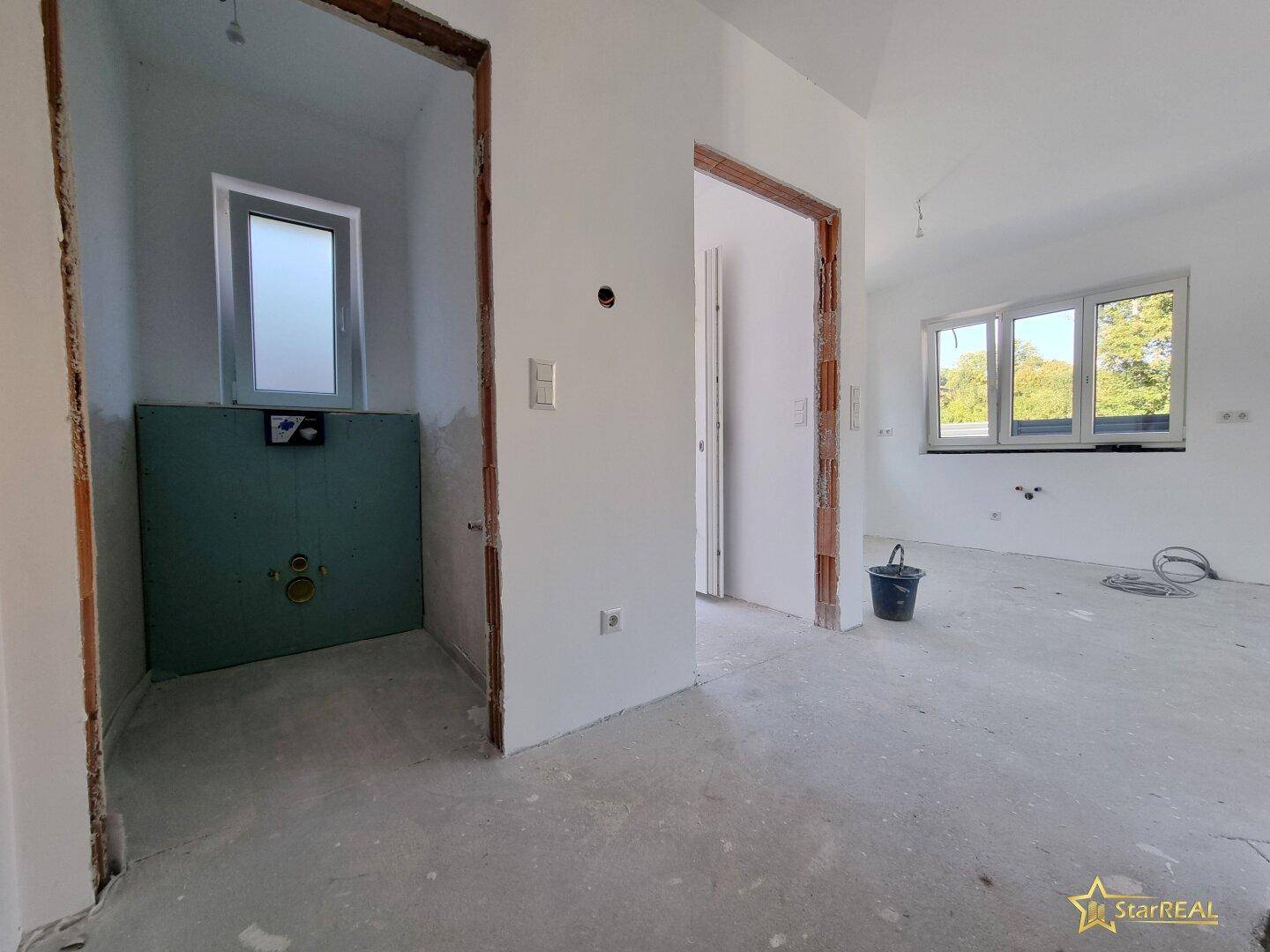 Innenraum im Rohbauzustand mit vorbereitetem Badbereich und Blick ins Grüne.