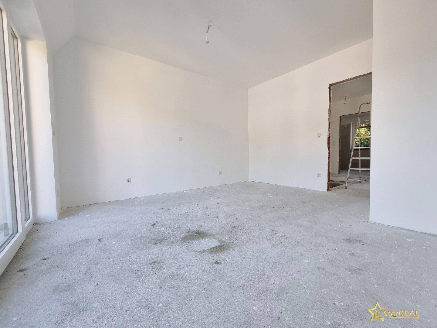 Raum im Rohbau mit viel Tageslicht und einer Leiter, die auf weitere Arbeiten hindeutet.