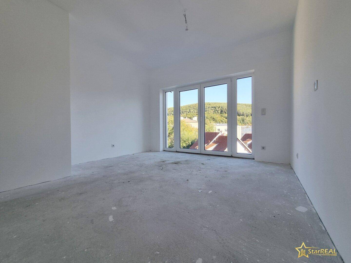 Heller Raum im Rohbau mit großen Fenstern und Blick auf die grüne Umgebung.