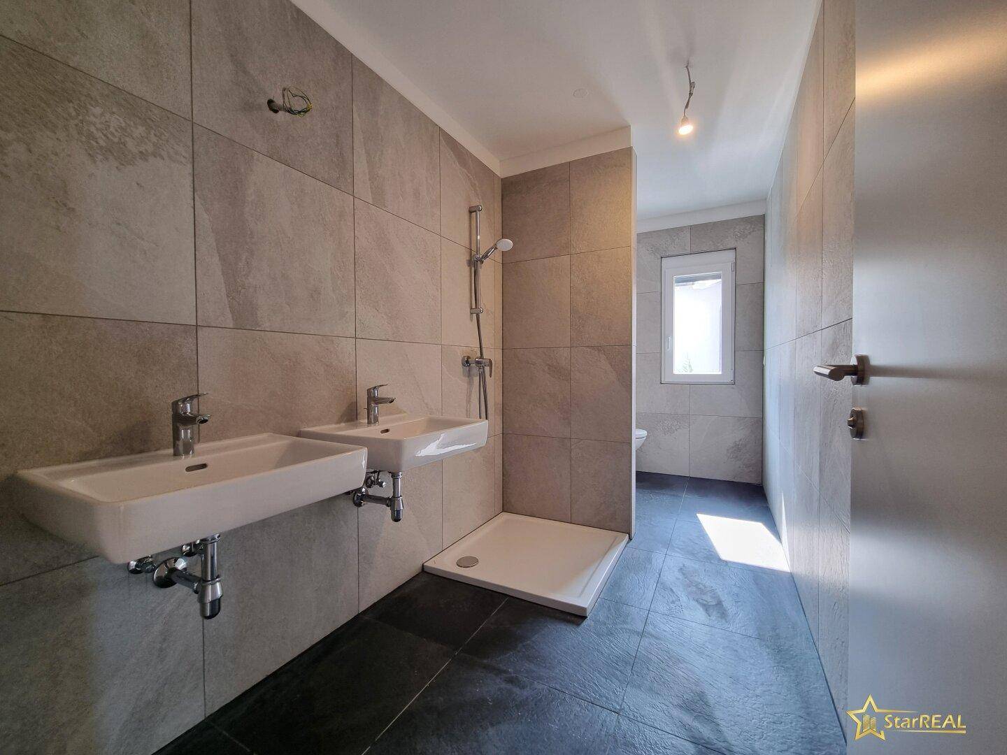Modernes Badezimmer mit Doppelwaschbecken, Duschbereich und grauen Fliesen.