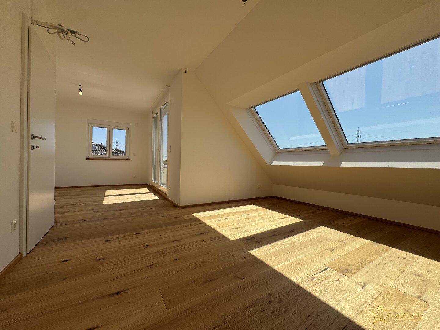 Helles Zimmer mit Holzboden, Dachschrägen und großen Fenstern, die viel Tageslicht hereinlassen.
