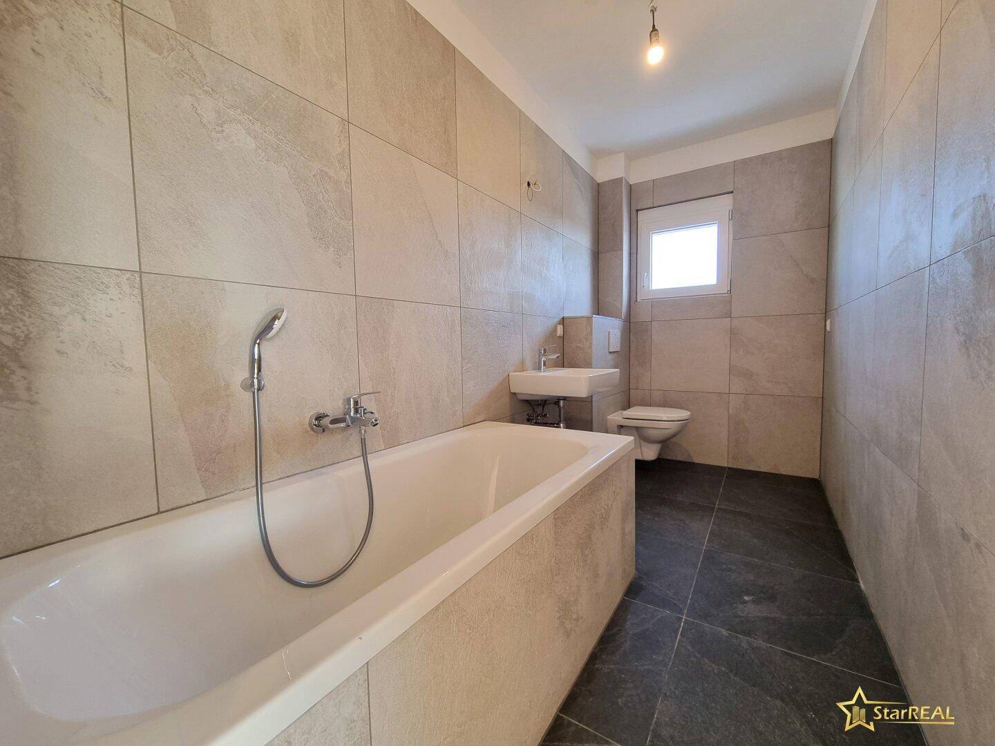 Modernes Badezimmer mit Badewanne, Waschbecken und WC, komplett gefliest in Grau.