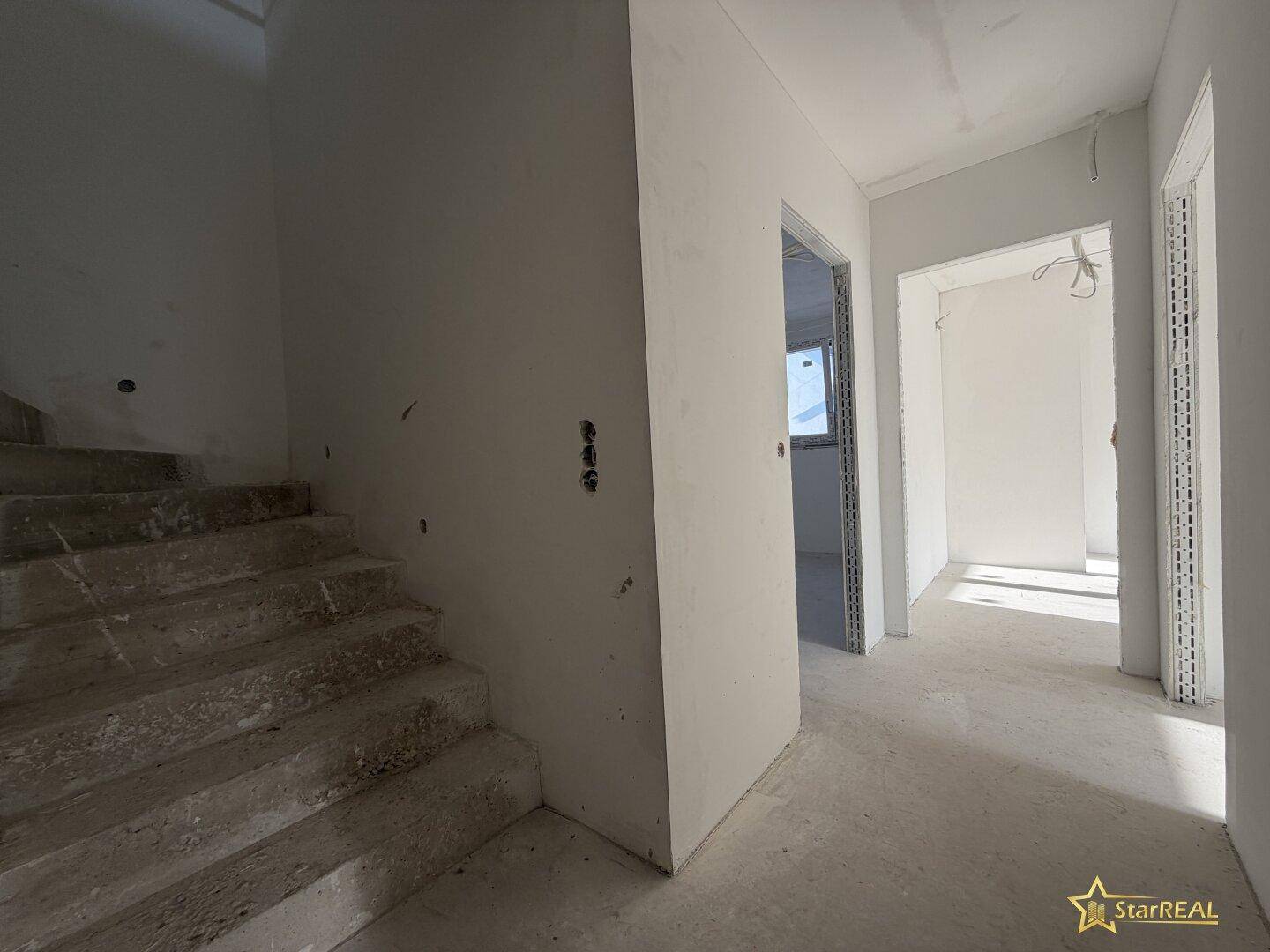 Flur im Rohbau mit Betontreppe und Zugang zu weiteren Räumen, bereit für den Ausbau.