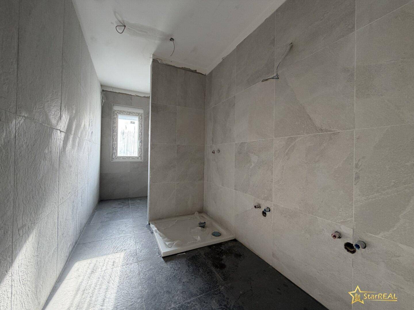 Badezimmer im Rohbau mit grauen Fliesen, Duschwanne und Fenster, bereit für die Installation.