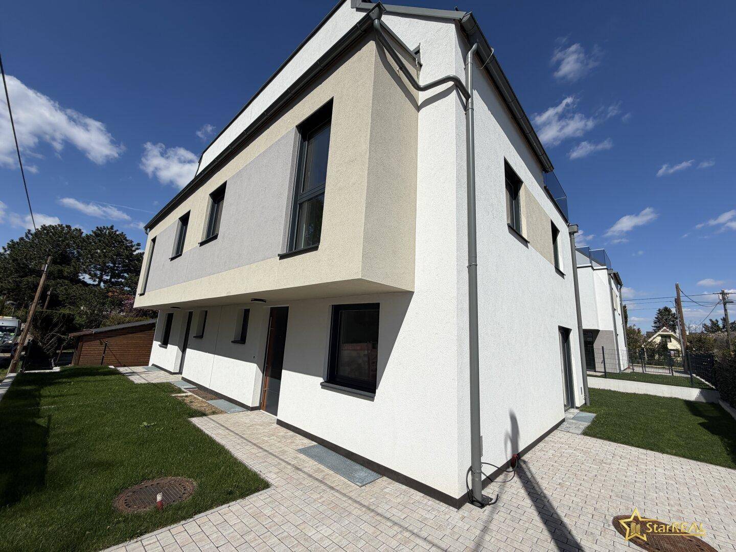 Seitenansicht eines modernen Neubaus mit heller Fassade und dunklen Fensterrahmen.