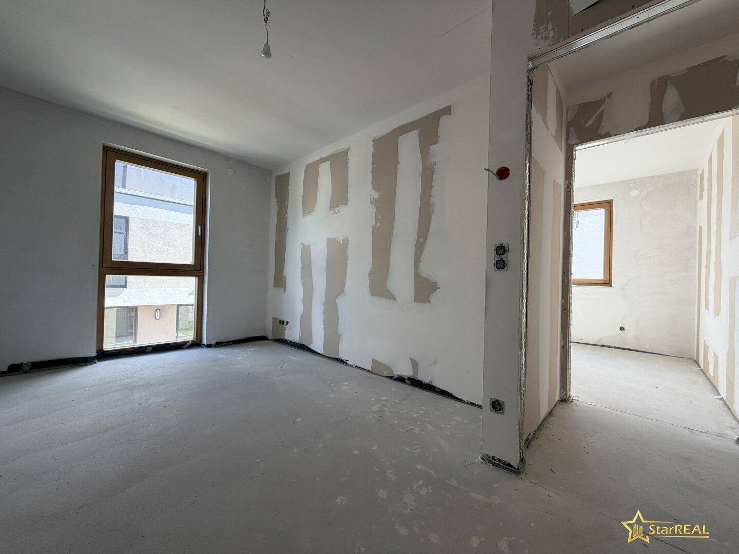 Lichtdurchfluteter Rohbau-Raum mit Fenster und Zugang zu einem weiteren Zimmer.