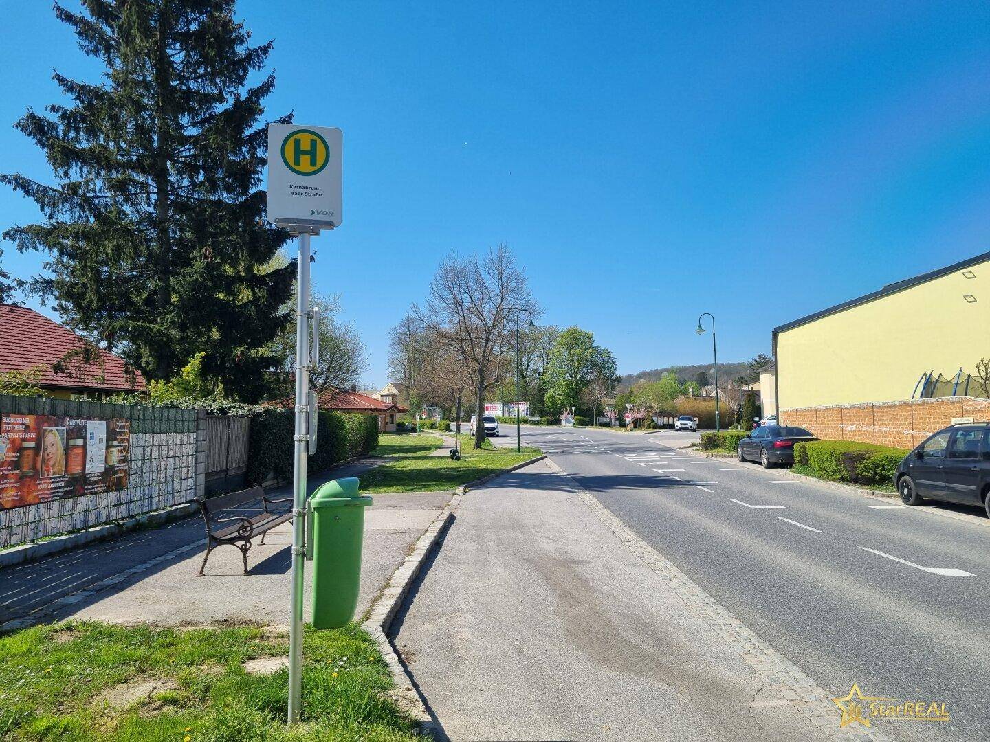 Bushaltestelle an einer ruhigen Straße mit Bäumen und Häusern im Hintergrund.
