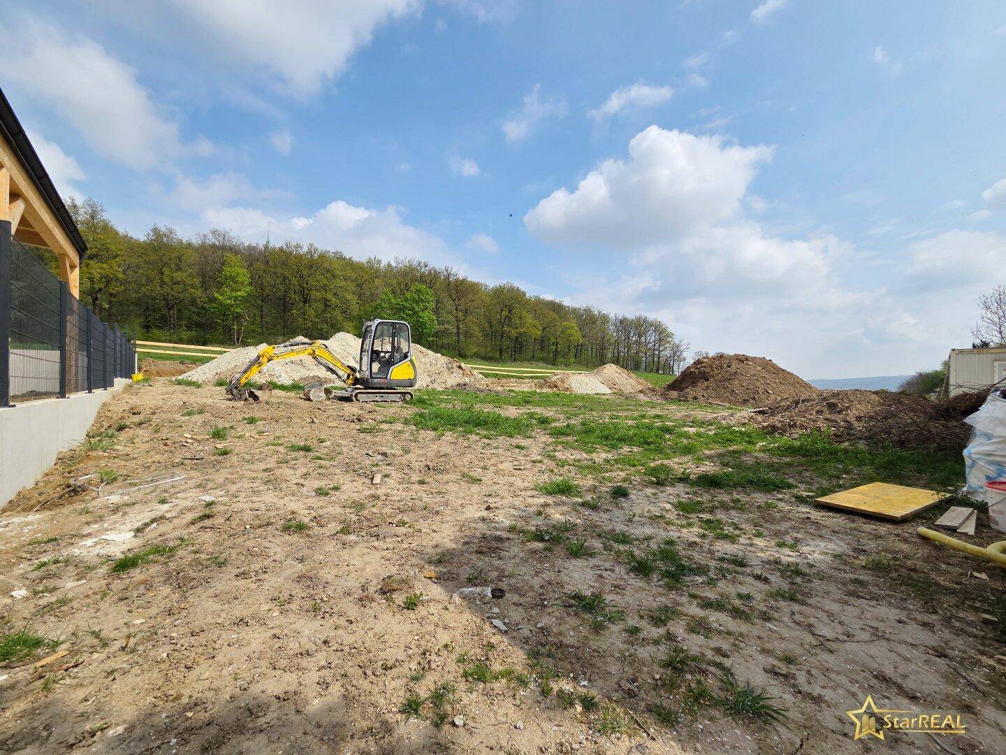 Baustelle mit Bagger und Erdhaufen, umgeben von grüner Landschaft.